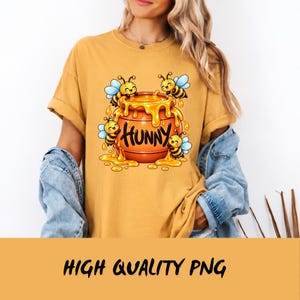 May include: A mustard yellow t-shirt featuring a graphic of a round orange honey pot with the word HUNNY written in black script. Three cartoon bees with blue wings are positioned around the pot, which is overflowing with yellow honey.