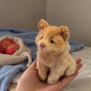 May include: A small, light tan, fuzzy plush animal figurine with rounded ears, a dark nose, and dark eyes. The creature has a soft, textured appearance and is depicted in a seated position, showcasing a compact size that fits within a palm.