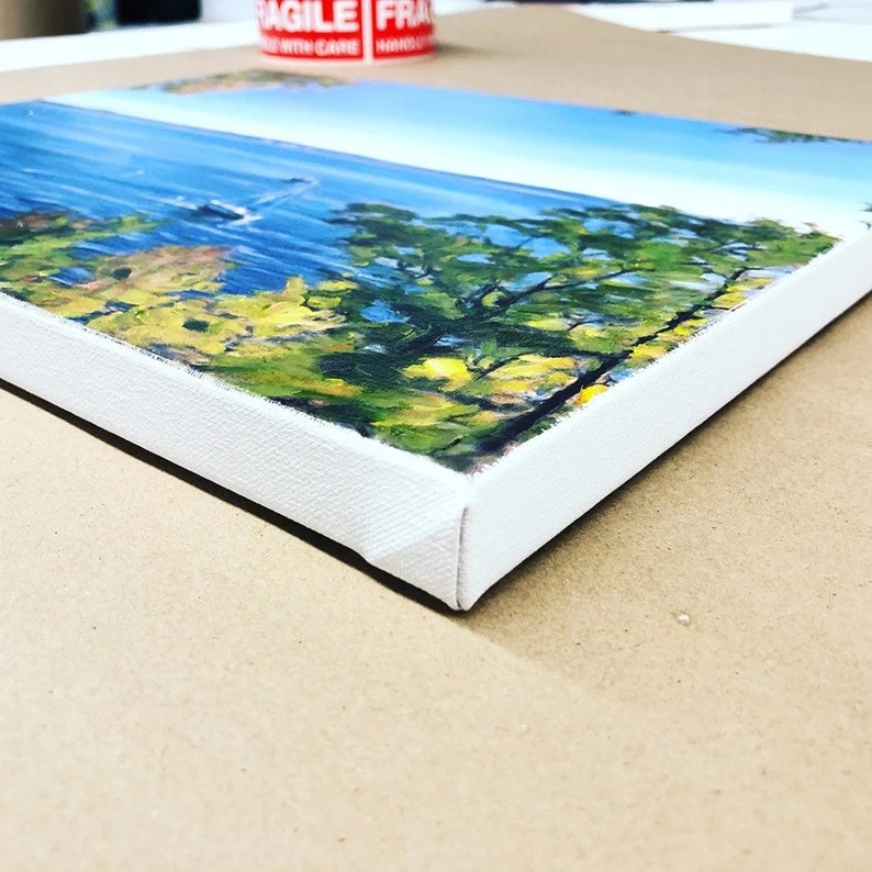 May include: Close-up of the edge of a canvas painting with a white frame. The painting depicts a landscape with trees and a blue body of water in the background.