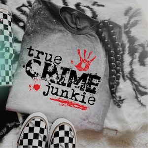 May include: A gray short-sleeve t-shirt with a bleached effect featuring the black text "true CRIME junkie" and a red handprint graphic. The design includes a red paint splatter and a red horizontal brush stroke accent below the text.