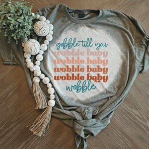 May include: A grey short-sleeved t-shirt with a bleached white circular center featuring the text "gobble till you wobble baby wobble baby wobble baby wobble baby wobble" in teal, pink, and orange. A wooden bead garland with tassels is included.