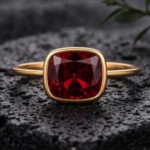 May include: A gold ring featuring a large, cushion-cut, deep red gemstone set in a smooth, polished bezel setting. The band is thin and circular, crafted from a warm-toned yellow metal that complements the vibrant, faceted red stone.