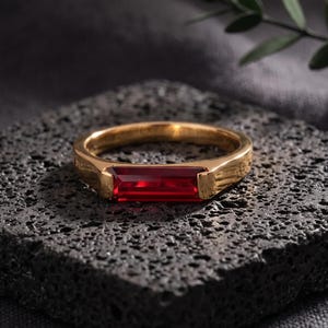 Op de afbeelding: Een goudkleurige ring met een enkele, rechthoekige, baguette-geslepen rode edelsteen die horizontaal in het midden van de band is gezet. De ring heeft een gladde, gepolijste metalen afwerking en een strak, lineair ontwerp dat de steen accentueert.