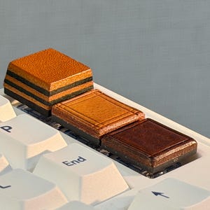 Puede incluir: Tres teclas cuadradas con superficies de cuero marrón texturizado están instaladas en un teclado de computadora blanco. Las teclas presentan distintos tonos de bronceado, marrón medio y marrón oscuro, con costuras visibles en los bordes.