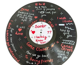 May include: A black vinyl record featuring hand-painted white and red lyrics and symbols. Text includes: 12 to 12, Dime, I wanna love you til the end of my life, Under the Mat, Sombr 77, I barely know her, Come Closer, Canal Street, and more.
