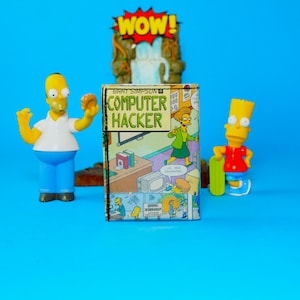 May include: A colorful image featuring three The Simpsons figurines and a comic book. The comic book cover reads "BART SIMPSON COMPUTER HACKER". Homer Simpson is holding a donut. Bart Simpson is holding a skateboard. The background is a blue surface.