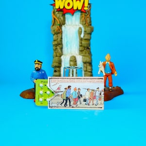 Könnte beinhalten: Eine skurrile Szene mit Miniaturfiguren und einem Comicstrip. Ein Wasserfall-Hintergrund mit einem "WOW"-Schild befindet sich hinter einem Glas. Eine Matrosenfigur, ein Comicstrip und eine Figur mit einem Hund befinden sich vor dem Wasserfall. Eine grün-weiße Pillenpackung ist ebenfalls vorhanden.