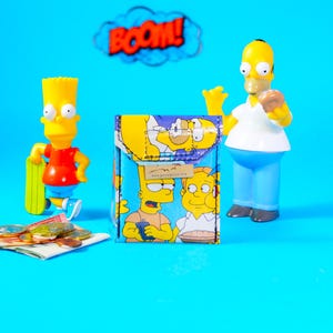 May include: A small, square wallet featuring a cartoon design of Bart and Homer Simpson, with a blue background. The wallet is surrounded by Bart and Homer Simpson figurines, coins, and a "BOOM!" speech bubble.
