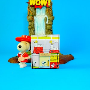 May include: A Snoopy-themed wallet with a comic strip design featuring Snoopy and Woodstock. The wallet is multi-colored with a black border. A Snoopy figurine in a red outfit is in front of the wallet. A waterfall and "WOW!" sign are in the background.