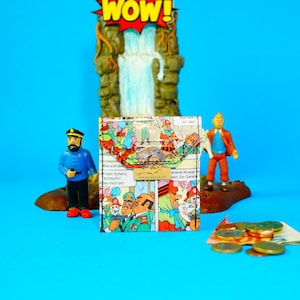 Könnte beinhalten: Eine farbenfrohe Geldbörse mit Comic-Panels, Sprechblasen und dem Wort "WOW!" in einem gelb-roten Ausbruch. Die Geldbörse wird mit Figuren und Euro-Münzen auf blauem Hintergrund präsentiert.