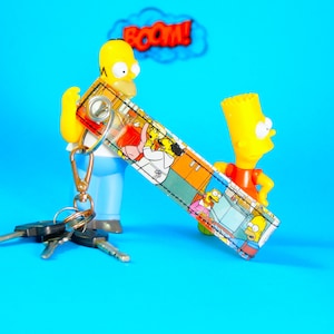 Może przedstawiać: Żółta postać z kreskówki trzyma brelok z kreskówkowym obrazkiem rodziny Simpsonów. Brelok ma srebrny metalowy pierścień i przymocowany czarny i srebrny klucz. Tło jest niebieskie.