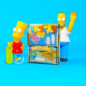 May include: A colorful wallet featuring comic strip panels with Bart Simpson illustrations. The wallet is displayed with Bart and Homer Simpson figurines on a bright blue background. The wallet has a small tag with a website address.