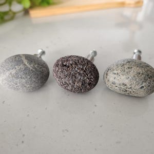 Può includere: Tre pomelli per armadietti con cime in pietra naturale in varie tonalità di grigio e nero. Il pomello a sinistra è liscio con una venatura bianca, quello centrale ha una consistenza scura e porosa, quello a destra è grigio maculato e ruvido.