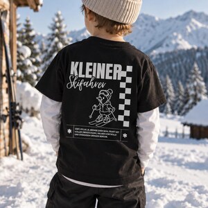 Könnte beinhalten: Ein schwarzes Kurzarm-T-Shirt mit dem weißen Schriftzug KLEINER Skifahrer, einer Grafik einer Ente auf Skiern, einem Schachbrettmuster und deutschem Text über einen Skifahrer, getragen über einem weißen Langarmshirt mit beiger Strickmütze.
