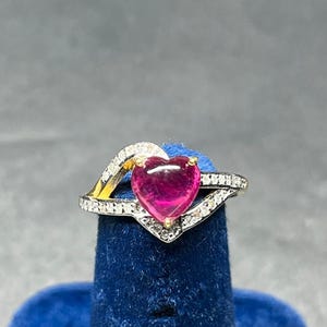 Pode incluir: Um anel com uma pedra preciosa rosa em forma de coração cravada em ouro amarelo, ladeada por duas bandas prateadas incrustadas com pequenas pedras claras que se curvam em torno do coração para criar um design assimétrico.