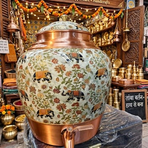 Puede incluir: Un gran dispensador de agua de cobre con cuerpo redondeado y tapa a juego. El exterior presenta una superficie de color crema decorada con un patrón repetitivo de elefantes azul oscuro con mantas naranjas, intercalados con motivos florales.