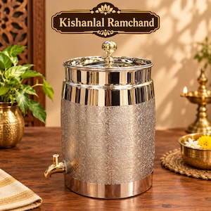 Puede incluir: Un dispensador de agua cilíndrico de acero inoxidable con acabado pulido, que presenta un intrincado patrón floral en relieve en el cuerpo, un grifo de metal dorado y una tapa a juego con un pequeño pomo esférico. El texto Kishanlal Ramchand es visible.