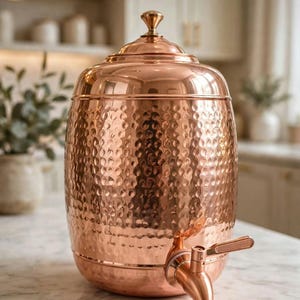 Könnte beinhalten: Ein Wasserbehälter aus gehämmertem Kupfer mit Deckel und Zapfhahn, der oben den Schriftzug Kishan Lal Ramchandra trägt. Das zylindrische Gefäß hat eine polierte metallische Oberfläche und einen abgerundeten Boden zur Flüssigkeitsaufbewahrung.