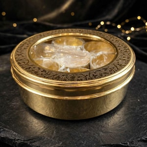 Könnte beinhalten: Ein runder, goldfarbener Metallbehälter mit einem durchsichtigen, kreisförmigen Deckel. Der Deckel hat einen dunklen, dekorativen Rand mit einem komplizierten Blumenmuster. Im Inneren sind mehrere kleine, goldfarbene Gegenstände in Klarsichtfolie zu sehen.