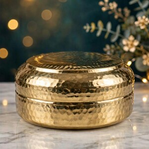 Puede incluir: Un recipiente redondo de metal dorado con tapa. La superficie exterior presenta una textura martillada en los lados y un patrón radial similar a un sol grabado en la parte superior de la tapa. El recipiente tiene un acabado pulido y brillante.