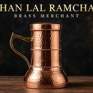 May include: A copper-colored metal mug with a hammered texture, a cylindrical body, and a flared base. The mug features a single handle and a flat lid. Above the mug, the text reads KISHAN LAL RAMCHAND BRASS MERCHANT in gold lettering.
