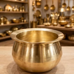 May include: A polished brass pot with a wide, rounded body and a flat, flared rim. The text KISHAN LAL RAMCHANDRA - Brass Merchants is visible at the top. The pot has a smooth, metallic finish and a deep, open interior, reflecting light on its surface.