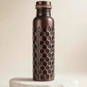 May include: A cylindrical copper water bottle featuring a dark, recessed honeycomb pattern across its body. The bottle has a smooth, rounded neck and a matching screw-top cap with a textured rim, showcasing a metallic finish with deep brown tones.