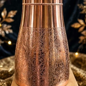 Peut inclure: Une bouteille d'eau en cuivre avec une partie supérieure lisse et une partie inférieure ornée d'un motif floral en relief. La bouteille possède un couvercle assorti et repose sur une boîte en carton avec le texte manuscrit 1580 et M0921E.