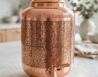 May include: A cylindrical copper water dispenser featuring an intricate floral engraving around the centre and a metal spigot at the base. The text "Kishan Lal Ramchand" is engraved near the top, and it includes a matching lid with a small knob.