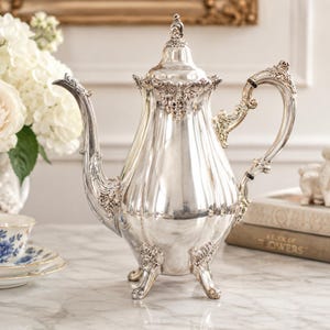 May include: A polished silver teapot featuring a fluted body, an ornate handle, a curved spout, and a decorative lid. The teapot stands on three detailed feet and displays intricate floral relief patterns around the base, handle, and spout.