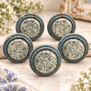 May include: Five round cabinet knobs featuring a cream-colored center with a blue floral pattern. The outer rim is painted in a distressed, weathered blue finish with visible brown patches, giving the hardware a worn, aged appearance.