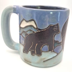 May include: A ceramic mug with a light blue and periwinkle glaze featuring a dark brown bear silhouette standing in front of a stylized mountain range. The mug has a thick, rounded handle and a smooth, glossy finish with visible dark outlines.