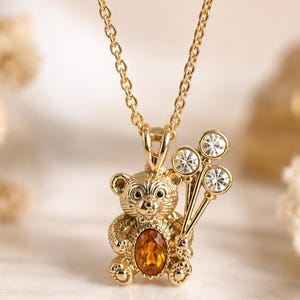 May include: A gold-toned necklace featuring a pendant shaped like a teddy bear. The bear has a textured surface, a central oval-shaped amber-colored gemstone on its chest, and holds three small, clear, round crystals arranged like balloons.