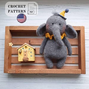 Puede incluir: Un elefante de ganchillo gris con un cuello de volantes amarillo y un gorro de fiesta amarillo a juego con un pompón azul. Junto a él hay un pequeño adorno de casa de madera amarilla con una campana. El texto dice CROCHET PATTERN PDF.