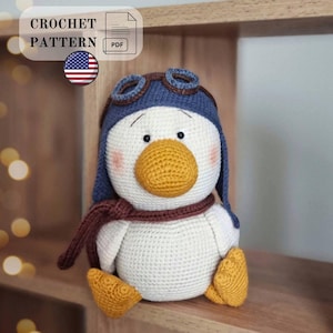 Pode incluir: Um padrão de crochê em PDF para um pato de pelúcia usando um chapéu de aviador azul com óculos marrons e um cachecol marrom. O pato tem corpo branco, bico amarelo, pés amarelos e bochechas rosadas. O texto CROCHET PATTERN PDF é visível.