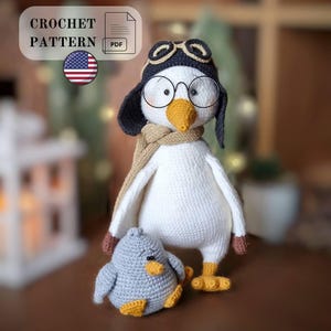 May include: A crochet pattern PDF featuring a white bird doll wearing a dark aviator hat with goggles, round wire-rimmed glasses, and a tan scarf, standing next to a smaller, round, light gray bird doll with a yellow beak and feet.
