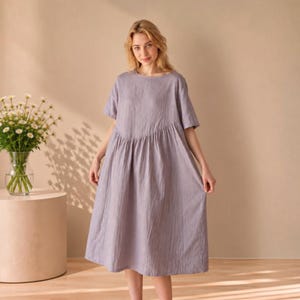 May include: A lavender midi dress featuring short sleeves, a round neckline, and a diagonal seam across the bodice that transitions into a gathered, flowing skirt. The garment is made of a textured fabric and falls to a mid-calf length.