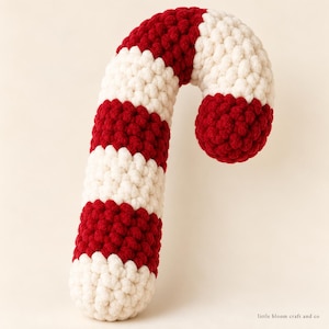 Puede incluir: Un adorno de bastón de caramelo tejido a ganchillo con rayas horizontales alternas de hilo rojo oscuro y blanco roto. La textura se crea con puntos densos y redondeados. El texto "little bloom craft and co" aparece en la esquina inferior derecha.