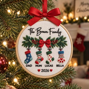 May include: A round embroidery hoop ornament featuring white fabric with the embroidered text The Brown Family. It displays a green pine garland with a red bow and four hanging stockings labelled DAD, MOM, LUCAS, and BELLA, with 2026 below.