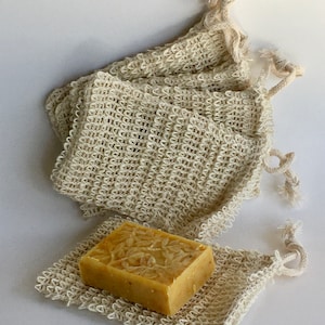 May include: A natural-coloured soap bar with a floral design sits on a matching woven soap saver. Several similar soap savers are stacked behind it. The soap and savers are made of a light beige material, creating a spa-like aesthetic.
