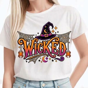 Puede incluir: Una camiseta blanca de manga corta con un estampado gráfico que muestra la palabra WICKED en letras con degradado naranja y morado. El diseño incluye un sombrero de bruja morado, telarañas, arañas naranjas, estrellas y una luna creciente.
