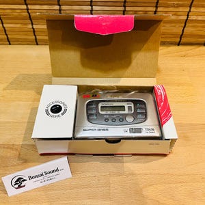 Pode incluir: Um leitor de cassetes portátil prateado com visor digital, rotulado como Super Bass e TX476, dentro de uma caixa de cartão aberta. Uma pequena caixa branca está marcada como Accessories Here. Inclui um cartão com o logótipo Bonsai Sound.