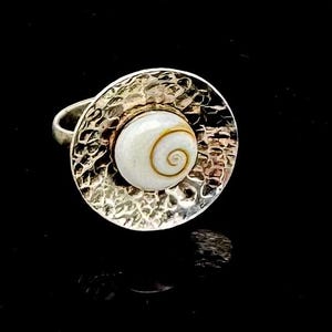 May include: A silver ring with a hammered texture and a large, round, white stone with a spiral design. The ring has a unique, eye-catching design, perfect for adding a touch of elegance to any outfit.
