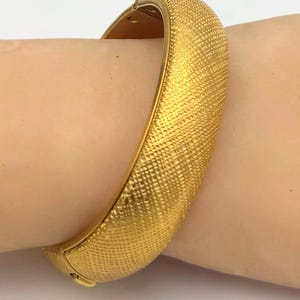May include: A gold-colored bangle bracelet with a textured surface. The bracelet is a solid band with a clasp closure. The surface has a repeating pattern of small squares. The bracelet is shown on a person's arm.