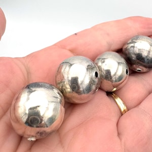 May include: Four spherical, silver-coloured beads with small holes. The beads are held in a hand, reflecting light. These beads are likely for jewellery making or other crafts. Each bead is approximately 2 cm in diameter.