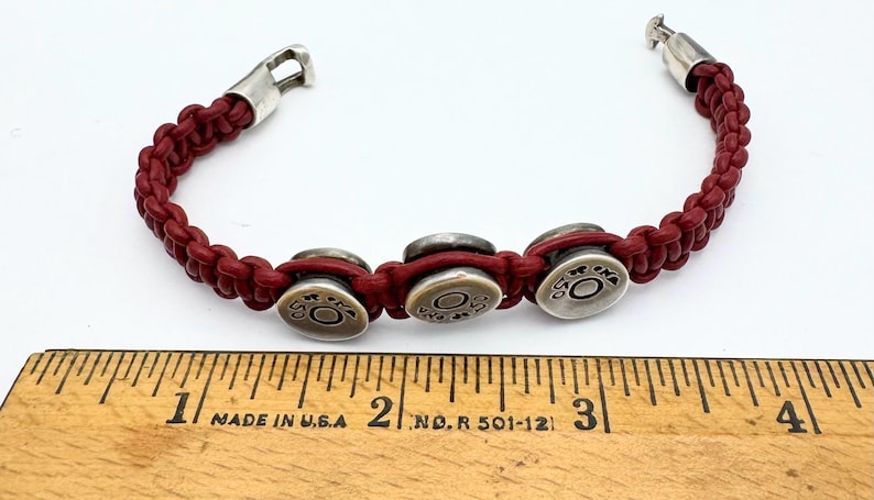 May include: A red leather bracelet with silver metal charms. The charms have the word "OSCAR" engraved on them.
