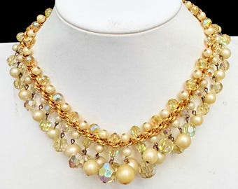 May include: A gold-tone chain necklace featuring a multi-layered fringe design composed of cream-colored round beads and faceted, iridescent light yellow crystal beads that dangle from the central metal chain links.