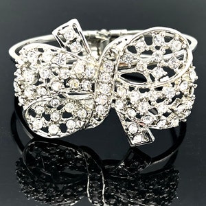 May include: Silver-toned bracelet featuring a bow design encrusted with numerous clear crystals. The bracelet has a hinged closure and reflects light, creating a sparkling effect. The bow is the focal point, adding a touch of elegance.