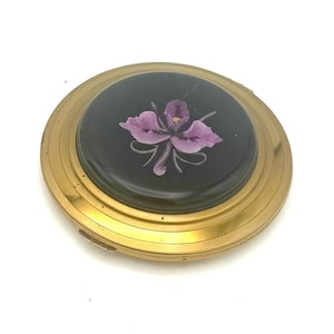 May include: A vintage compact with a gold-toned metal casing. The center features a black circular design with a purple flower illustration. The compact has a hinged opening and a reflective surface.