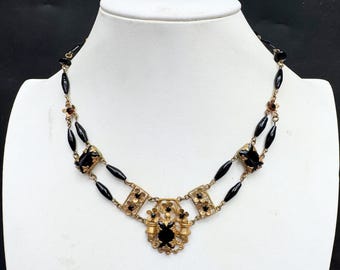 May include: A gold-tone metal necklace featuring a double-strand design with elongated black beads, small floral accents, and a central pendant. The piece is adorned with faceted black stones set within ornate, filigree-style metal frames.